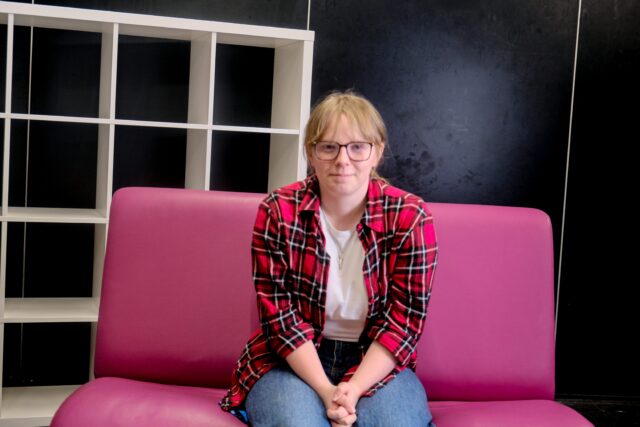 Katie sat on pink sofa, smiling with hands clasped.