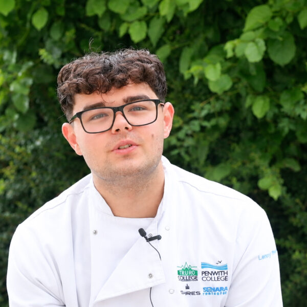 Lenny A student in a Truro and Penwith College chef’s jacket and glasses sits outdoors in front of green foliage, wearing a microphone for an interview