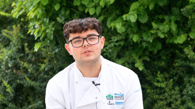Lenny A student in a Truro and Penwith College chef’s jacket and glasses sits outdoors in front of green foliage, wearing a microphone for an interview