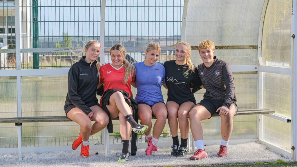 5 girls rugby players, seated in dugout smiling