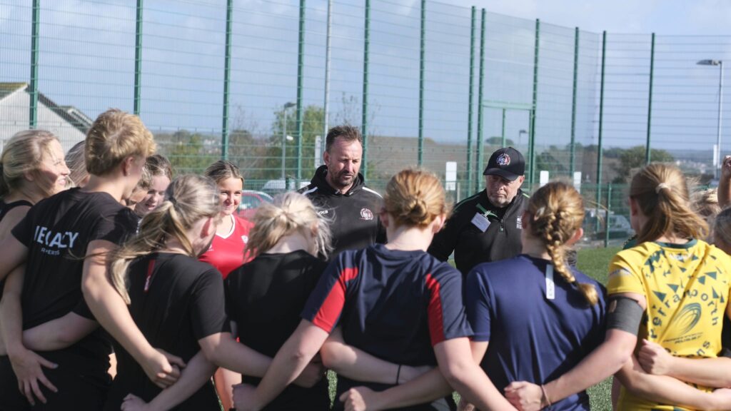 Two male coaches depicted in background, delivering a talk to group of girls stood with arm interlinked, backs to camera