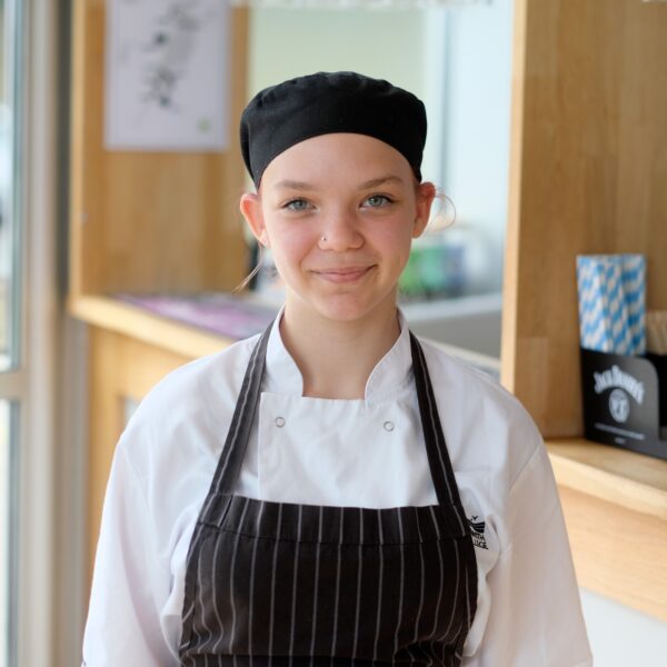 Picture of young girl smiling in black chef hat and apron, in front of a bar.
