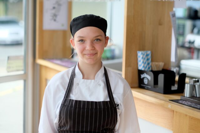 Picture of young girl smiling in black chef hat and apron, in front of a bar.